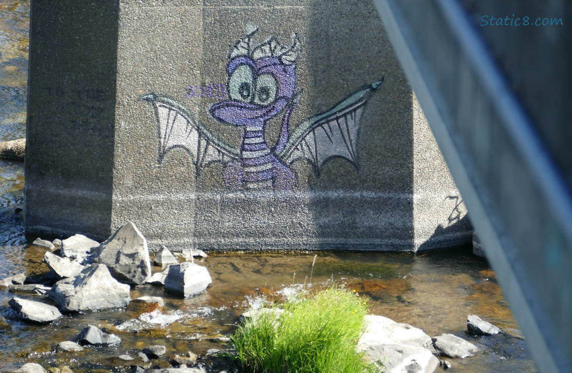 Cute Dragon graffiti on bridge support, water and rocks beneath